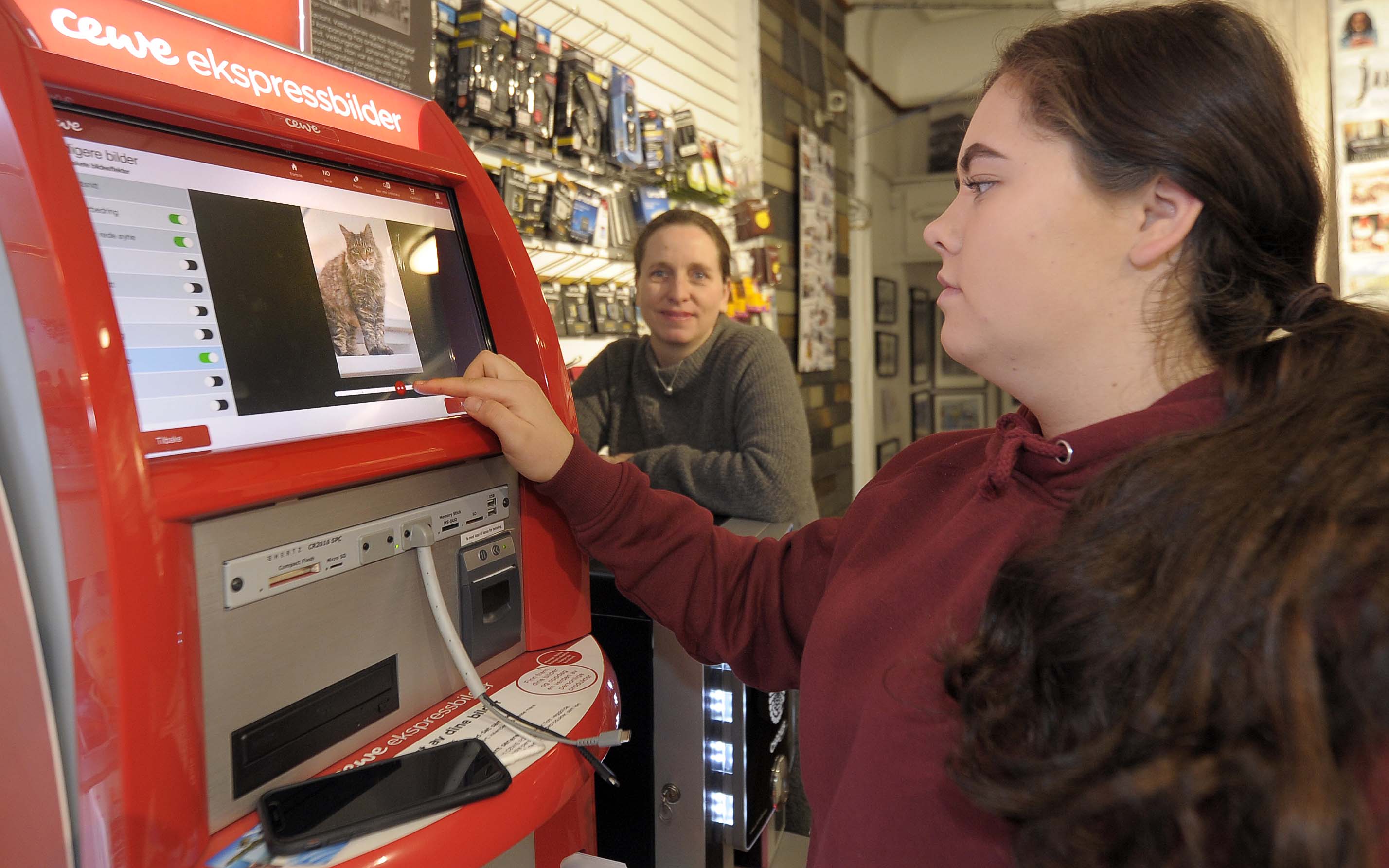 Celina Vartdal (t.h) har funnet katten på mobilen, klar til å printe ut. -Vi har fire slike automater i butikken, forteller Marit Brunstad.