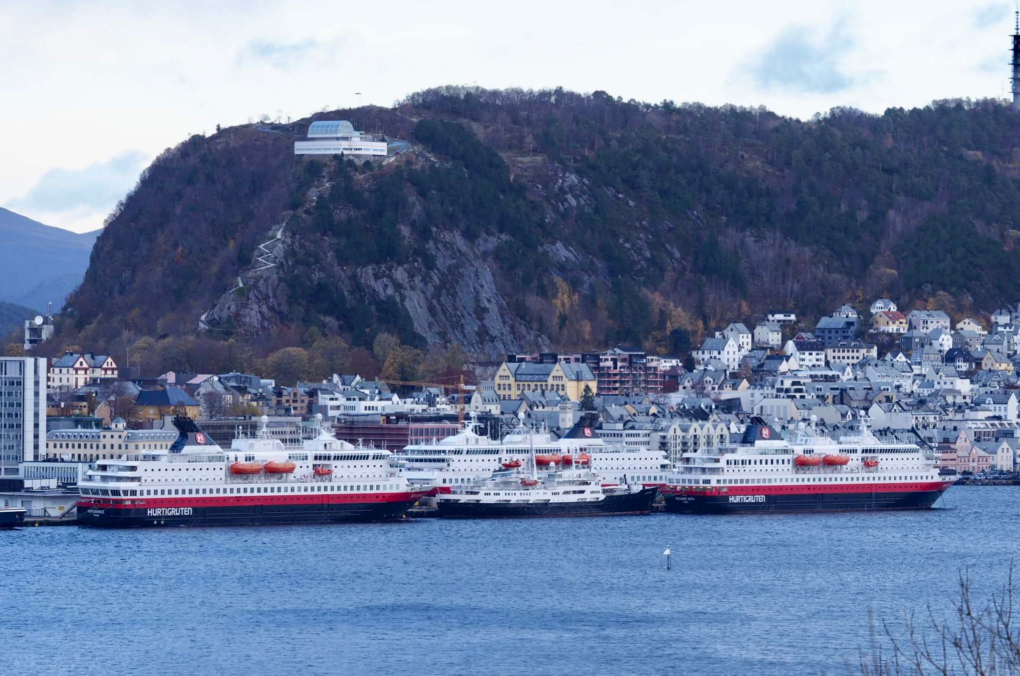 En rekke Hurtigruter til kai i Ålesund sentrum. Bak ser du Fjellstua. En tur de ansatte om bord gjerne tar daglig!